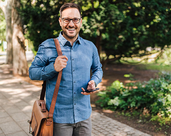 Portrait of smiling male executive walking on his way to work. Young professional commuter with smart phone is carrying laptop bag.