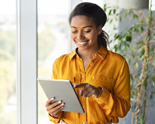 Happy black female entrepreneur using tablet standing near window at modern coworking office, chatting with client. Focused lady browsing internet